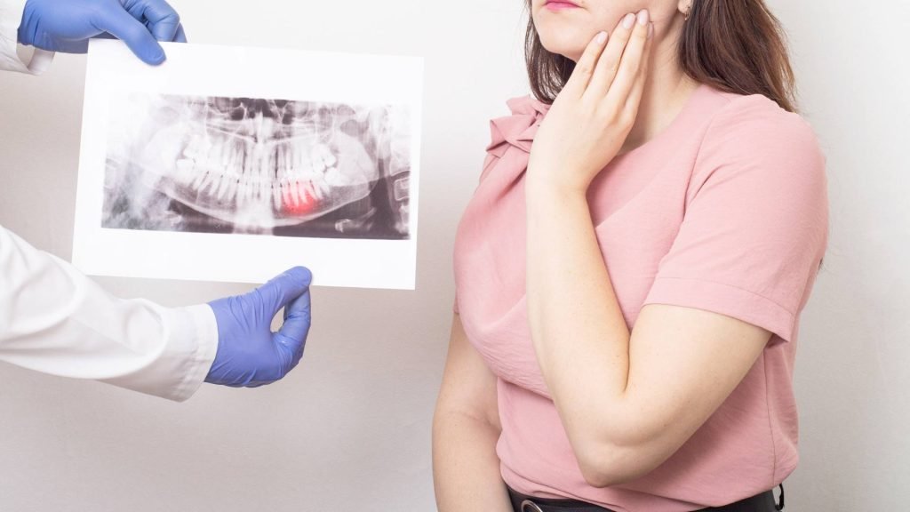 A woman sits in a doctor's office holding her jaw while a doctor holds up an X-Ray, demonstrating the jaw surgery process.