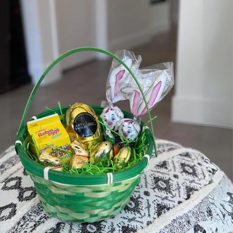 A vibrant Easter basket filled with assorted treats, including chocolate eggs and a yellow box of Peeps, sits on a white lace tablecloth. This festive arrangement is perfect for Philadelphia orthodontists and dentists to consider for Easter Basket Deliveries